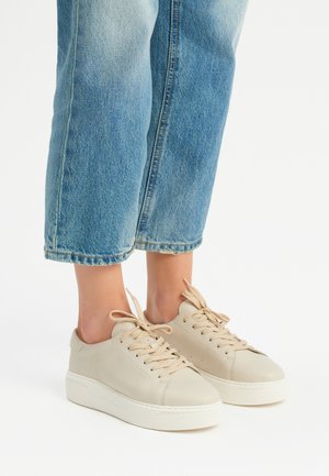Beżowe skórzane sneakersy o teksturowanym wykończeniu, okrągłym nosku i kremowej gumowej podeszwie. Posiadają beżowe sznurówki i płaski fason. Noszone z jasnoniebieskimi dżinsami.