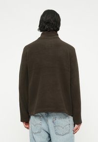 Sudadera de forro polar marrón con cuello alto y mangas largas. Presenta una superficie texturizada y un detalle de parche cuadrado en la parte posterior.
