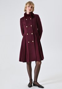 Bordeaux wollen jurk met een geplooide rok, dubbelgeknoopte gouden knopen en een nepbont kraag. Gladde textuur, lange mouwen, knie-lengte.