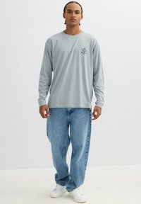 Camiseta de manga larga gris claro con un pequeño gráfico en el pecho, combinada con jeans holgados de color azul claro y zapatillas blancas.