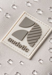 Šedé tkaninové pozadí se vzorovanou texturou. Výrazná šedá guma logo nášivka s nápisem "audetic" na spodní straně, lemovaná bílou.