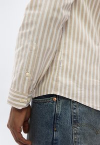 Gros plan d'une personne portant une chemise à rayures verticales beige et blanc avec des poignets boutonnés et un jean denim bleu avec des coutures visibles.