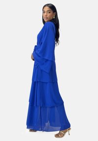 Blå lagdelt maxikjole med lange ærmer, talje med snøring og gennemsigtigt stof; båret med gyldne remsandaler og dråbeøreringe.