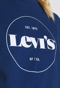 Námořnický modrý svetr s bílým kruhovým logem Levi's, včetně textu "EST. 1873" a "SF | CA." na přední části v oblasti hrudi.