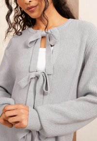 Cardigan tricotat de culoare gri deschis, cu guler rotund, detalii cu fundiță în față și manșete cu nervuri, având o textură moale.