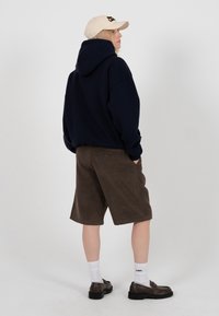 Personne portant un sweat à capuche marine, une casquette beige, un short marron mi-genoux, des chaussettes blanches et des mocassins foncés, debout les mains dans les poches, de profil.