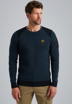 Dunkelblauer Strickpullover mit gerippten Akzenten an den Schultern und Bündchen. Mit einem kleinen gelben Logo-Patch auf der Brust. Strukturierter Stoff.