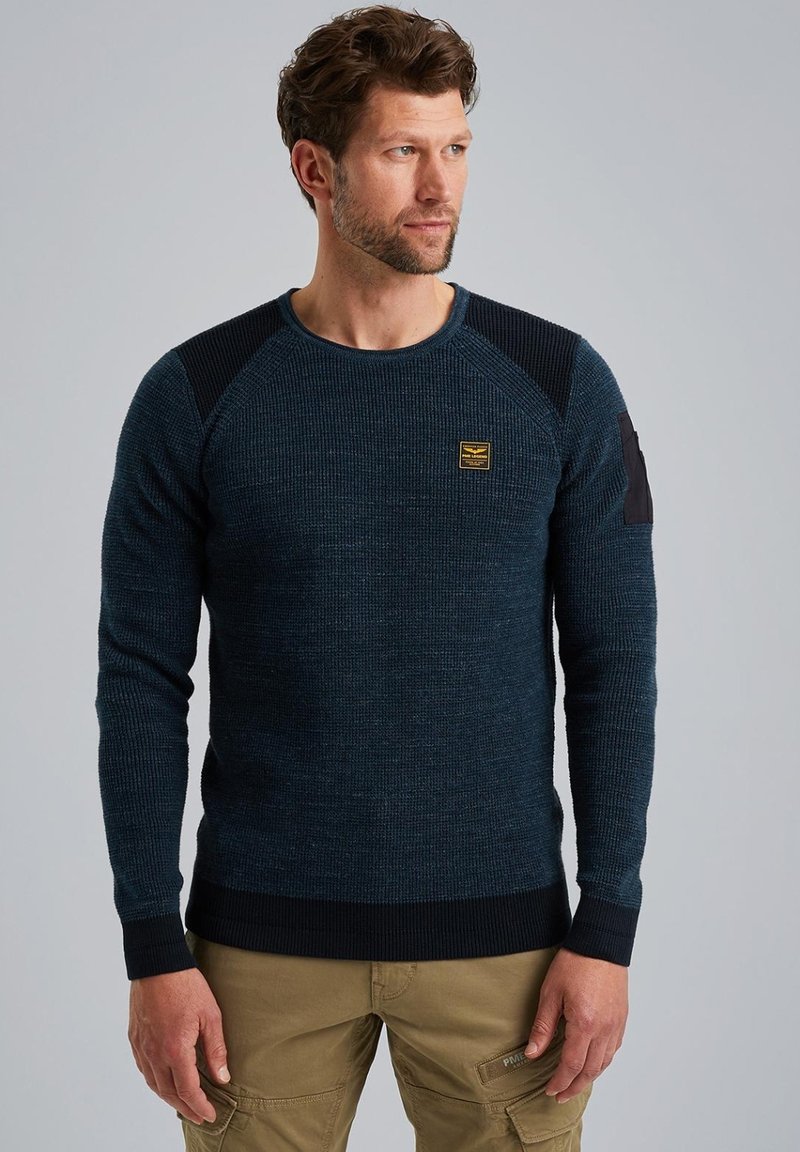 Dunkelblauer Strickpullover mit gerippten Akzenten an den Schultern und Bündchen. Mit einem kleinen gelben Logo-Patch auf der Brust. Strukturierter Stoff.