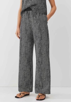 Frau trägt lockere, schwarz-weiß gemusterte Hosen mit weitem Bein und ein passendes ärmelloses Oberteil, steht mit den Händen in den Taschen und schwarzen Sandalen.