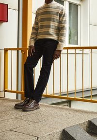 Randig tröja i beige och grått, svarta jeans och bruna snörade stövlar med en texturerad sula, som står mot ett gult räcke.