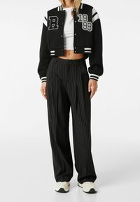 Černá zkrácená varsity bunda s bílými pruhy a nápisy, kombinovaná s černými kalhotami s jemným proužkem. Boty jsou světlé s černými podrážkami.