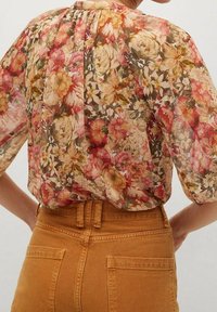 Transparante bloemenblouse met opgestoken mouwen, in een hoge taille mosterdkleurige broek met achterzakken gestoken.