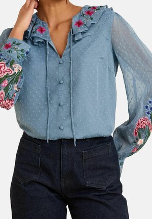 Lyseblå tekstureret bluse med stofbetrukne knapper, blomsterbroderi på kraven og gennemsigtige ærmer, kombineret med mørke høj-taljede jeans.