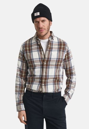 Man in een zwarte beanie, witte T-shirt, bruine en witte geruite overhemd met knopen, en donkere broek, staande met een hand in de zak.