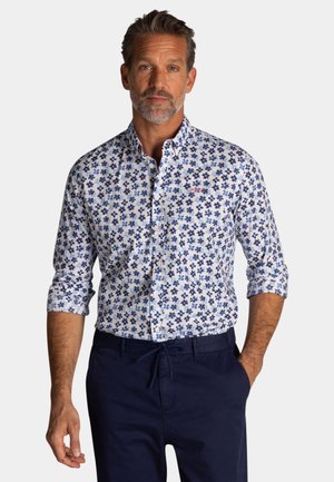 Man met kort haar en baard, draagt een wit overhemd met blauwe bloemenprint en donkerblauwe broek, handen in de zakken.