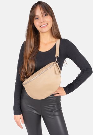 Junge Frau mit langen braunen Haaren lächelt, trägt ein schwarzes Oberteil, schwarze Lederhose und eine beige Umhängetasche mit Reißverschluss.