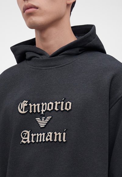 Jeune personne portant un sweat à capuche gris foncé avec "Emporio Armani" et un logo d'aigle rayé brodé sur la poitrine.