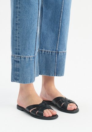 Pieds chaussés de sandales à enfiler en cuir tressé noir, associés à un jean en denim bleu raccourci sur un fond blanc.