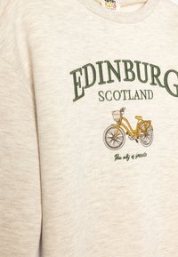 Sudadera color crema con texto bordado en verde "EDIMBURGO ESCOTIA", que presenta un gráfico de bicicleta amarilla y una tela suave y texturizada.
