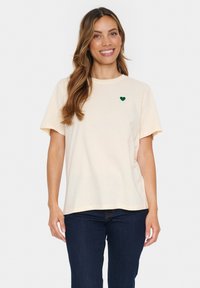 Tricou din bumbac de culoare crem, cu guler rotund și mâneci scurte. Are un mic inima verde brodată în zona stângă a pieptului.