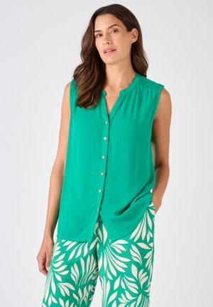 Mujer con blusa verde sin mangas con botones y pantalones verdes y blancos con estampado, de pie con una mano en el bolsillo frente a un fondo liso.