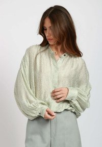 Blusa verde chiaro semi-trasparente con un motivo a trama, vestibilità comoda, colletto alto e chiusura con bottoni. Presenta maniche voluminose e polsini arricciati.