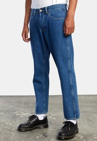 Vaqueros de mezclilla azules con corte recto, diseño de cinco bolsillos y costuras en contraste. Llevados con zapatos negros y calcetines blancos sobre una superficie de concreto.