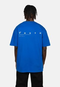 Kék, túlméretezett pamut póló, fehér "LOST YOUTH" felirattal és grafikai elemekkel a hátsó részen. Rövid ujjú, laza szabású.