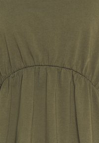 Olivengrønt stof med rynkede stingdetaljer, der skaber bløde folder og tekstur i et horisontalt bånd på tøjet.