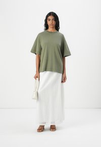 Olijfgroene oversized t-shirt, witte maxi rok van kant met oogjesdetails, gecombineerd met tan gekruiste sandalen en een kleine witte handtas.