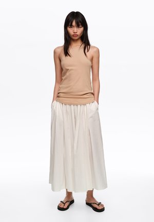 Mujer con top sin mangas beige, falda larga blanca con bolsillos y sandalias planas negras, posando frente a un fondo blanco liso.