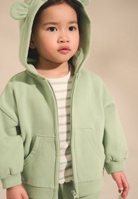 Ljusgrön zip-up hoodie med björnöron, mjuk textur och framfickor, lager-på-lager över en vit och grönrandig skjorta.