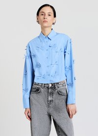 Chemise bleue à manches longues avec des nœuds en tissu surélevés en guise d'ornement. Assortie à un jean en denim gris taille haute.
