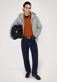 Šedá mikina na zip, oranžové polo tričko, tmavě modré kalhoty, černé rukavice, drží černou tašku. Hnědé boty s hladkým povrchem. Jednoduchý a moderní design.