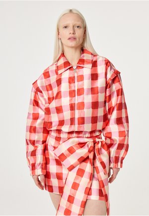 Vrouw draagt een rood-roze geruit jack met een ritssluiting en oversized mouwen, gecombineerd met een bijpassende rok die ter hoogte van de taille is vastgebonden met een grote strik.