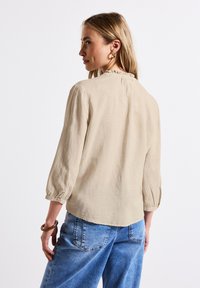 Beige linne-top med trekvartslånga ärmar och en volanghalsringning. Texturerat tyg med en avslappnad passform, kombinerat med högmidjade blå jeans.