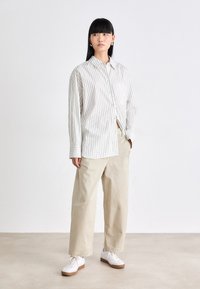 Giovane donna con capelli neri lisci che indossa una camicia bianca a righe sottili, pantaloni beige a gamba larga e sneaker bianche, in piedi su uno sfondo neutro.