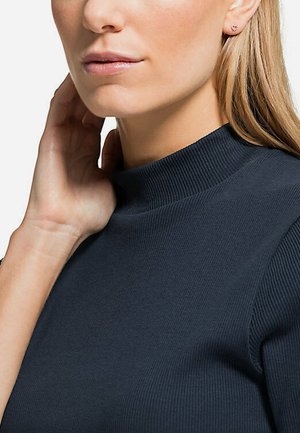 Maglietta a collo alto a coste blu navy con maniche lunghe, caratterizzata da un tessuto testurizzato e una silhouette aderente. Sono visibili orecchini a bottone discreti.