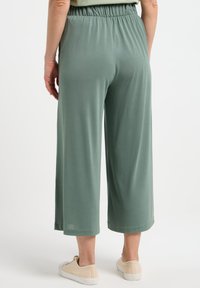 Pantalons mi-hauts à jambes larges en tissu vert doux et atténué. Dotés d'une taille élastique et d'une texture lisse. Portés avec des chaussures de couleur claire.