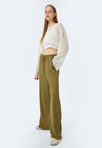Maglione corto in pizzo bianco con maniche lunghe e ampie abbinato a pantaloni verdi oliva, larghi e testurizzati. Sfondo chiaro, palette di colori neutri.