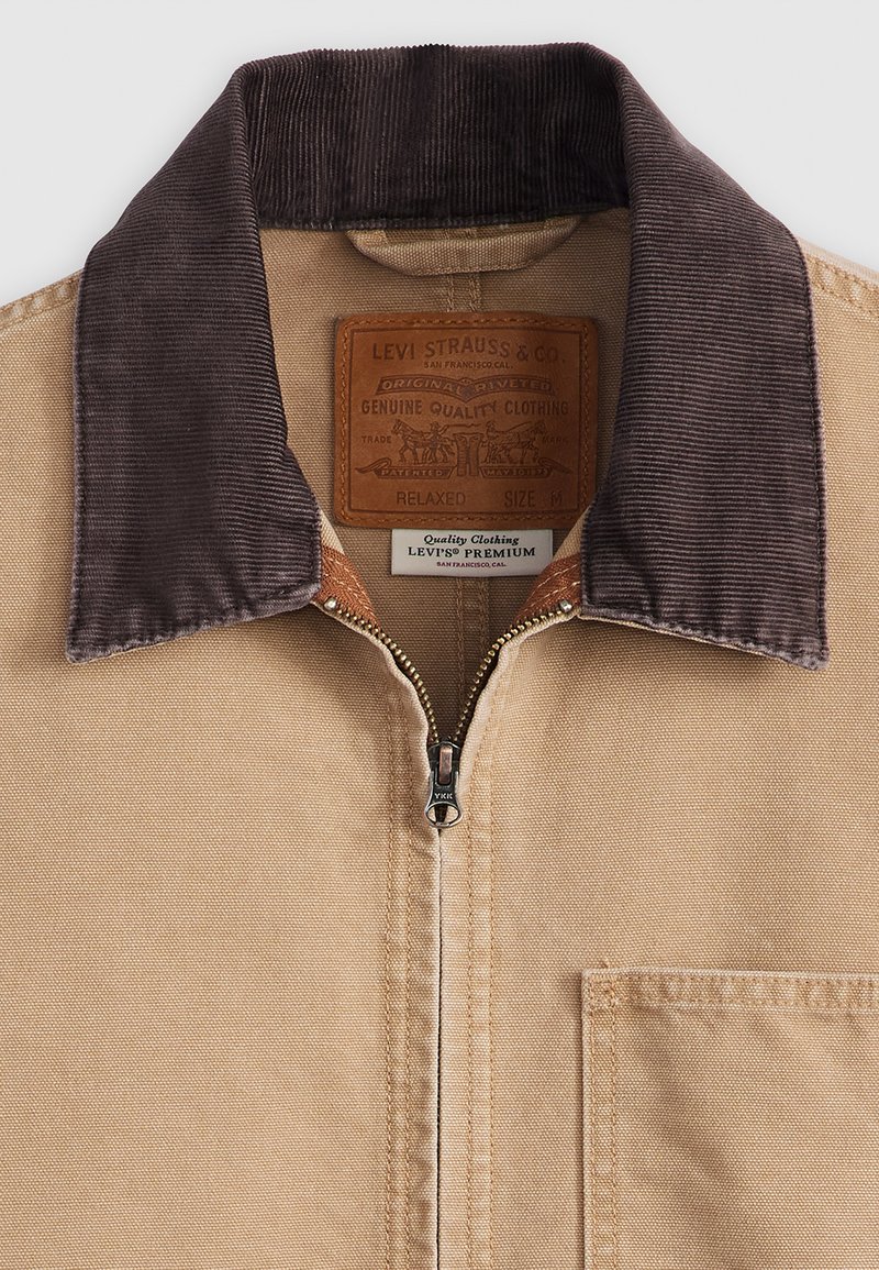 Chaqueta tipo zip en tono beige con un collar de pana marrón, cremallera de metal y un bolsillo en el pecho. Presenta una etiqueta de cuero con información de la marca.