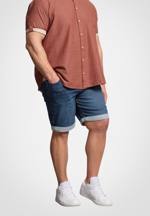 Mand iført rustfarvet kortærmet skjorte med knapper, op-rullede cowboyshorts og hvide sneakers, stående med hænderne i lommerne.