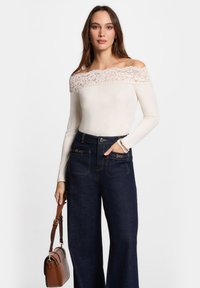 Cremfarbener Off-Shoulder-Top mit Spitzenbesatz und gerippter Textur, kombiniert mit dunkelblauen lässigen Jeanshosen mit weitem Bein und seitlichen Reißverschlusstaschen.