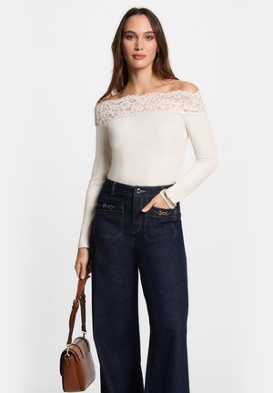 Top crema off-the-shoulder con rifinitura in pizzo, texture a coste, abbinato a pantaloni in denim a gamba larga blu scuro con tasche frontali con zip.