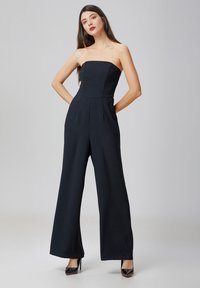 Combinaison noire sans bretelles avec jambes larges, corsage ajusté et plis subtils à la taille. Tissu lisse avec une finition élégante, associée à des talons hauts.