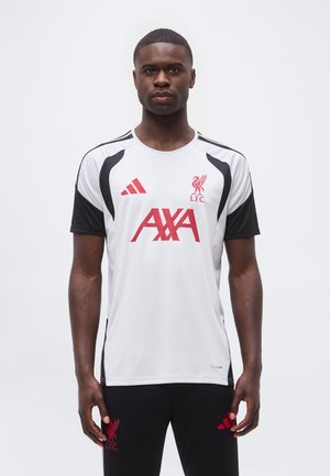 Homme portant un maillot d'entraînement blanc et noir du Liverpool FC avec des logos rouges AXA et Adidas, debout devant un fond blanc uni.