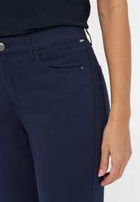 Navyblauwe vrouwenbroek van een gladde, rekbare stof met zijzakken, discreet beslag en een aansluitend ontwerp bij de taille en heupen.