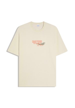 Bézs rövid ujjú póló kerek nyakkivágással, narancssárga szöveggel: „A BÉKE OTT KEZDŐDIK, AMIKOR MAGAMRA FIGYELEK” és egy kis virág grafikával.