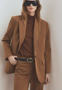 Bruine op maat gemaakte blazer over een bruine gebreide trui, gecombineerd met bijpassende broek. Zwarte riem met gouden gesp. Model draagt overgrote zonnebril.