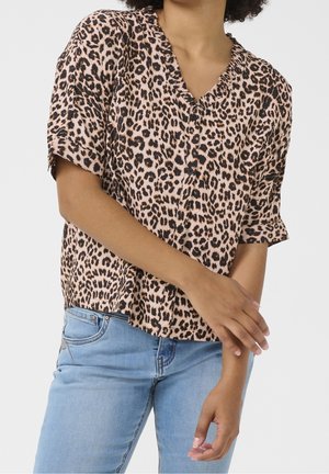 Frau trägt eine kurzärmelige Bluse mit Leopardenmuster und Knöpfen, kombiniert mit hellblauen Jeans, steht mit verschränkten Händen auf der Taille.